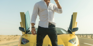 Quiet Luxury. Foto homem branco com camisa branca e calça jeans furada no joelho em frente a um carro de luxo