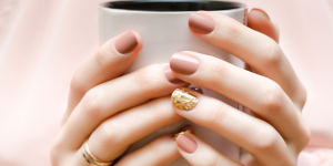 Nail Arts elegantes para mulheres de 30 anos. Foto aparece mãos segurando uma xícara branca. Unhas trazem esmalte nude e detalhes em dourado.