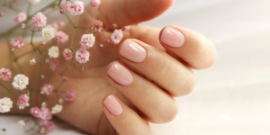 unhas curtas delicadas. Foto mostra mão sobre flores e com a unha pintada de nude